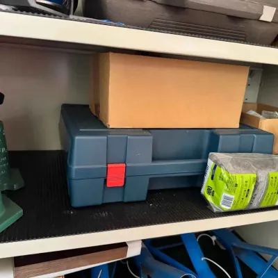 Sale Photo Thumbnail #126: Assorted workshop items on a shelf, including a blue plastic tool case with a red latch, a cardboard box, a package of steel wool, and green metal bench vises.