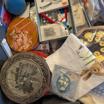 Sale Photo Thumbnail #160: A plastic storage bin filled with vintage sewing supplies, including decorative tins, snap fasteners, a hole punch, a blue pincushion, and embroidered linens.