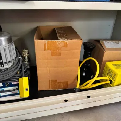 Sale Photo Thumbnail #128: Porta Power hydraulic pump unit with corded remote, shown alongside cardboard boxes and a yellow foot pedal switch on a metal shelf.