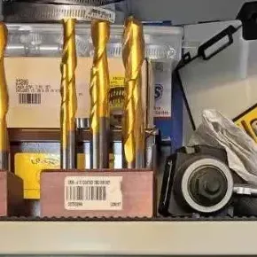 Sale Photo Thumbnail #111: Assorted gold-coated drill bits in wooden stands, alongside various boxed tools and hardware items on a shelf.