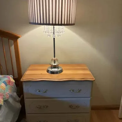 Sale Photo Thumbnail #185: Three-drawer nightstand with a natural wood top and painted gray base, paired with a modern table lamp featuring a striped drum shade and hanging crystal accents.