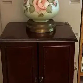 Sale Photo Thumbnail #184: Vintage Hurricane lamp with hand-painted pink roses on a green glass body, resting on a dark wood two-door cabinet with a lower drawer.