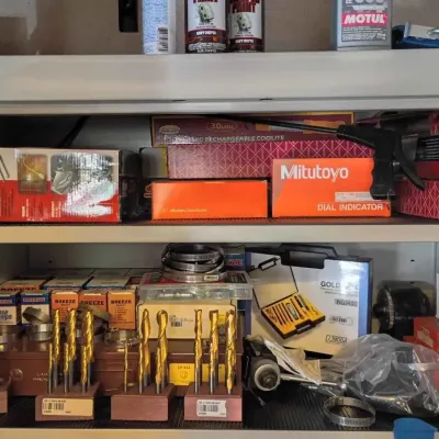 Sale Photo Thumbnail #110: Assorted workshop tools and equipment, including boxed Mitutoyo dial indicators, a Ridgid SeeSnake, various drill bit sets, and hose clamps on metal shelving.