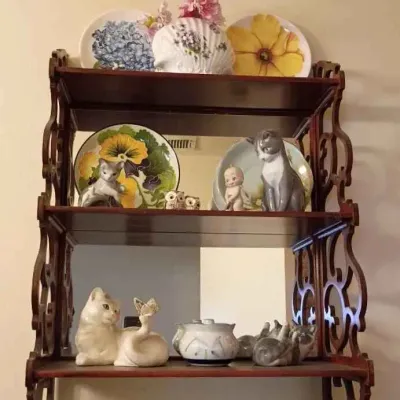 Sale Photo Thumbnail #145: Three-tier wooden wall shelf with ornate scrollwork sides, displaying a collection of decorative floral plates and ceramic cat figurines.