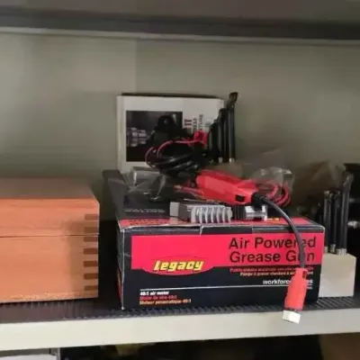 Sale Photo Thumbnail #131: Legacy air powered grease gun in its original box, shown alongside a wooden storage chest and a metal lathe chuck on a workshop shelf.
