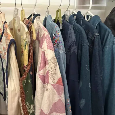 Sale Photo Thumbnail #90: Assortment of women's clothing hanging in a closet, featuring quilted vests, patterned jackets, and several denim shirts.