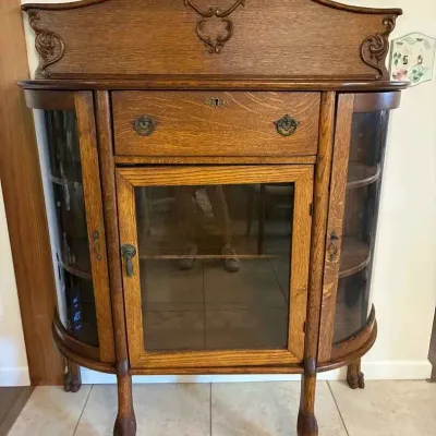 Sale Photo Thumbnail #13: Antique tiger oak parlor cabinet featuring curved glass side doors, a central glass door, a single drawer, and claw-and-ball feet, topped with an ornate carved backsplash.