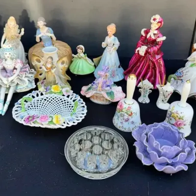Sale Photo Thumbnail #157: Collection of vintage porcelain figurines, floral bells, a woven ceramic basket, and decorative glassware displayed on a black surface.