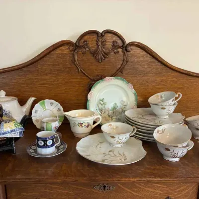 Sale Photo Thumbnail #159: Assorted vintage porcelain teacups, saucers, plates, and a teapot displayed on a carved wooden sideboard.