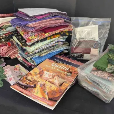 Sale Photo Thumbnail #34: Assorted quilting supplies, including stacks of colorful fabric remnants, pre-cut shapes, a plastic storage bin, and quilting pattern books.