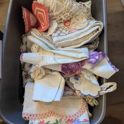 Sale Photo Thumbnail #161: A plastic storage bin filled with vintage linens, including crocheted doilies, embroidered dresser scarves, and lace-trimmed table runners.