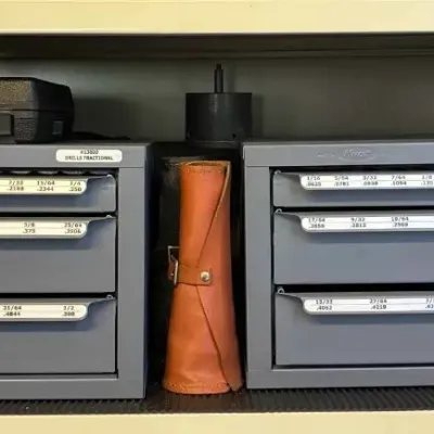 Sale Photo Thumbnail #116: Two Huot fractional drill bit dispensers with labeled drawers, shown alongside a leather tool roll and a black plastic storage case on a shelf.