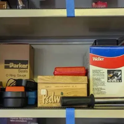 Sale Photo Thumbnail #106: A shelf of workshop tools and supplies, including a Binks paint spray gun, a Weller WES51 soldering station, a Parker Seals O-ring gauge, and various other hardware items.