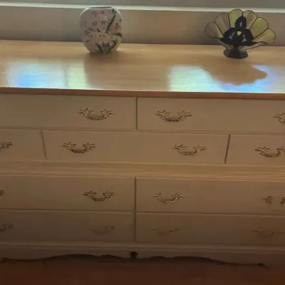 Sale Photo Thumbnail #186: White dresser with a light wood top and ornate gold-tone drawer pulls, displaying a small floral vase and a decorative fan-shaped stained glass piece.