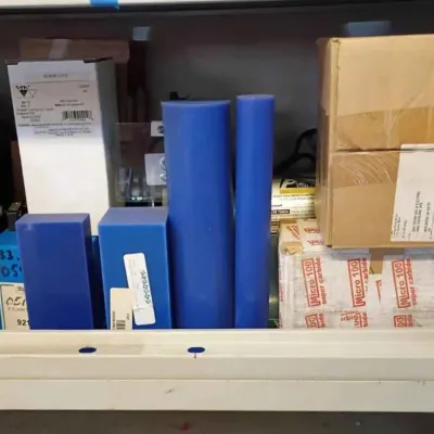 Sale Photo Thumbnail #115: Assorted industrial tooling and materials, including blue plastic stock blocks and cylinders, boxed Power Lock components, and Micro 100 super carbide tooling boxes stored on a metal shelf.