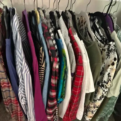 Sale Photo Thumbnail #89: Assorted women's clothing hanging on a closet rack, including plaid flannel shirts, striped blouses, and patterned tops.