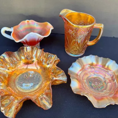 Sale Photo Thumbnail #141: Four-piece collection of vintage marigold carnival glass, including a pitcher with geometric patterns, a ruffled bowl with a starburst center, a floral-patterned ruffled dish, and a small handled nappy.