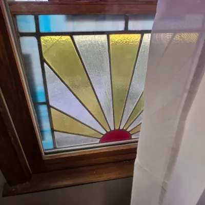 Sale Photo Thumbnail #143: Vintage stained glass window panel featuring a vibrant sunburst design in yellow, red, and blue tones, set within a wooden frame.