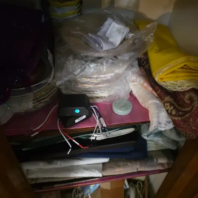 Sale Photo Thumbnail #100: Assorted household items, including folded fabrics, linens, wrapped plates, and a small electronic component with wires, stored on shelves inside a wooden cabinet.
