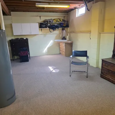 Sale Photo Thumbnail #185: Unfinished basement room featuring a vintage wooden dresser with an attached mirror, a black leather director's chair, and various storage furniture pieces.