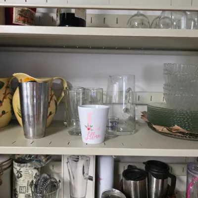 Sale Photo Thumbnail #149: Assorted kitchenware and barware on metal shelves, including a stainless steel cocktail shaker, ceramic pitchers, glassware, and a personalized "Lillian" mug.