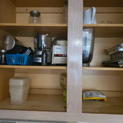 Sale Photo Thumbnail #126: Assorted kitchen appliances and storage items, including a Cuisinart food processor, an Osterizer blender, plastic food containers, and baking pans, arranged on wooden cabinet shelves.