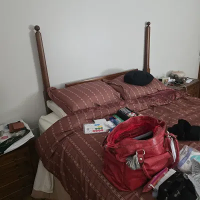 Sale Photo Thumbnail #122: Wooden four-poster bed frame with a reddish-brown patterned comforter and matching pillows, shown with various personal items and a red handbag resting on top.