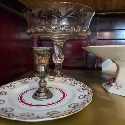 Sale Photo Thumbnail #24: Assorted vintage tableware including a tarnished silver-plated candlestick on a patterned porcelain plate, a clear etched glass compote, and a white ceramic pedestal dish.