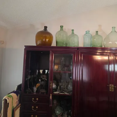 Sale Photo Thumbnail #5: Collection of vintage glass carboys and demijohn bottles in amber, green, and aqua tones, displayed atop a dark cherry wood china cabinet.