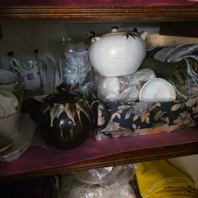 Sale Photo Thumbnail #99: Assorted vintage teapots, teacups, and saucers, including a brown drip-glaze teapot and a white ceramic teapot with bamboo motif, displayed on a wooden shelf.