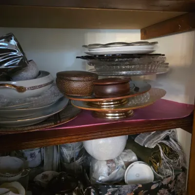 Sale Photo Thumbnail #98: Assorted vintage tableware and serving pieces, including glass platters, ceramic bowls, and wooden lidded boxes, arranged on wooden cabinet shelves.