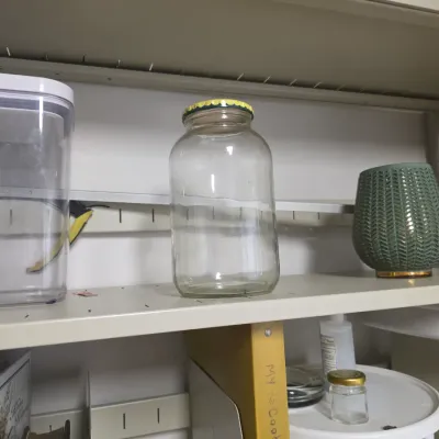 Sale Photo Thumbnail #136: Assorted household items on a shelf, including a large clear glass jar with a yellow lid, a green textured ceramic vase, and a clear plastic storage container.