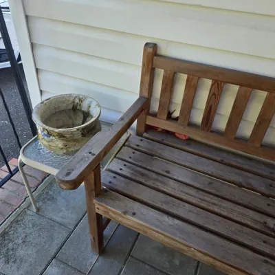 Sale Photo Thumbnail #215: Weathered wooden garden bench with a slatted seat and backrest, shown alongside a small glass-top side table holding a ceramic planter.