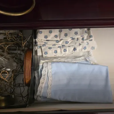 Sale Photo Thumbnail #19: Assorted vintage linens, including a blue pillowcase with lace trim and a floral patterned sheet, stored in a dresser drawer alongside metal chains and plastic stands.
