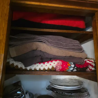 Sale Photo Thumbnail #97: Wooden cabinet shelves containing folded towels, a crocheted blanket, glass plates, and small decorative containers.