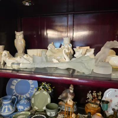 Sale Photo Thumbnail #31: Collection of vintage porcelain and glass collectibles, including white ceramic owls, a frosted glass figurine, and blue and green Wedgwood Jasperware pieces displayed on cabinet shelves.