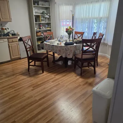 Sale Photo Thumbnail #125: Wooden dining set featuring an oval table with a floral tablecloth and five matching X-back chairs with brown upholstered seats, positioned in a dining area with wood-look flooring.