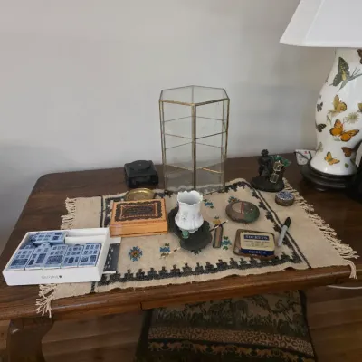 Sale Photo Thumbnail #81: Assorted tabletop decor on a wooden desk, including a butterfly-motif ceramic lamp, a hexagonal brass and glass display case, a small woven runner, and various vintage trinket boxes.
