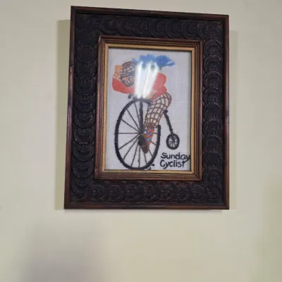 Sale Photo Thumbnail #208: Vintage framed needlepoint artwork titled "Sunday Cyclist," depicting a figure on a penny-farthing bicycle, housed in a heavily carved dark wood frame.