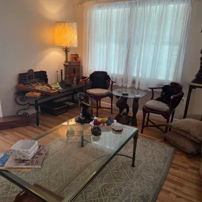 Sale Photo Thumbnail #48: Eclectic living room furniture and decor, featuring a glass-top coffee table, a pair of wooden armchairs, a vintage floor lamp, and various decorative sculptures.