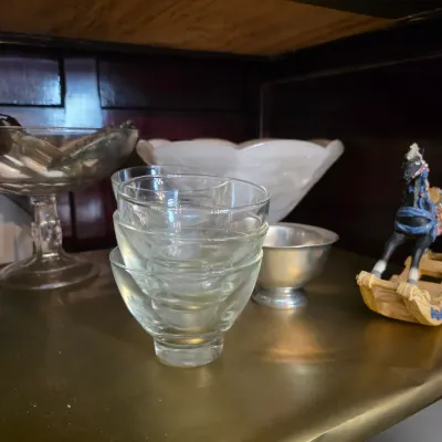 Sale Photo Thumbnail #23: Assorted glassware and decorative items, including a stack of clear glass cups, a pedestal bowl, a small metal dish, and a miniature rocking horse figurine.