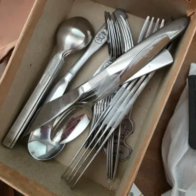 Sale Photo Thumbnail #111: Assorted stainless steel flatware, including spoons, forks, and knives, stored in a cardboard box.