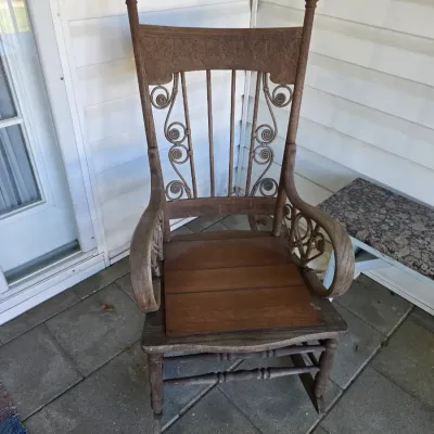 Sale Photo Thumbnail #213: Antique wooden rocking chair featuring ornate scrollwork on the backrest and under the armrests, showing some wear and weathering.