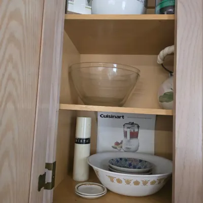 Sale Photo Thumbnail #129: Assorted kitchenware in a wooden cabinet, including a vintage Pyrex Butterfly Gold mixing bowl, a clear glass bowl, a Misto olive oil sprayer, and a white ceramic bowl with a black mushroom pattern.