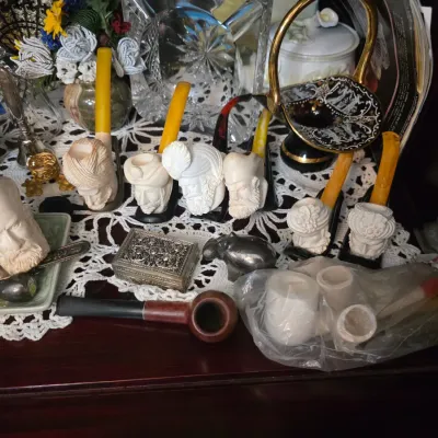 Sale Photo Thumbnail #38: Collection of vintage meerschaum smoking pipes carved with detailed faces and figures, displayed alongside a classic briar pipe, a small silver-toned trinket box, and various decorative glassware.
