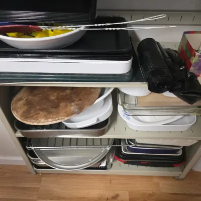 Sale Photo Thumbnail #151: Assorted kitchenware and baking dishes, including a pizza stone, glass casseroles, metal roasting pans, and plastic trays, organized on a metal shelving unit.