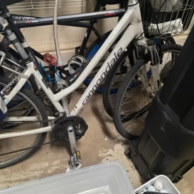 Sale Photo Thumbnail #161: White Cannondale hybrid bicycle with front suspension, showing some wear and dirt from use, parked next to other bikes in a garage.