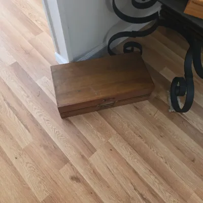 Sale Photo Thumbnail #70: Vintage rectangular wooden storage box with a metal latch, resting on a light wood floor next to a wrought iron table leg.