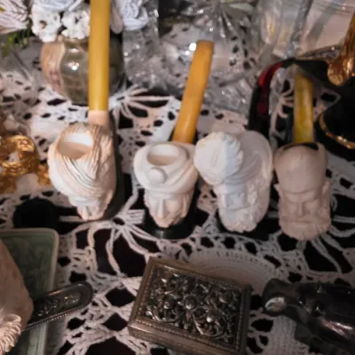 Sale Photo Thumbnail #39: Collection of vintage meerschaum smoking pipes with intricately carved character heads, displayed alongside a small silver-tone metal box on a white lace tablecloth.