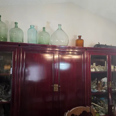 Sale Photo Thumbnail #6: Collection of vintage glass demijohns and bottles in various shades of green and amber, displayed atop a large mahogany-finished china cabinet.